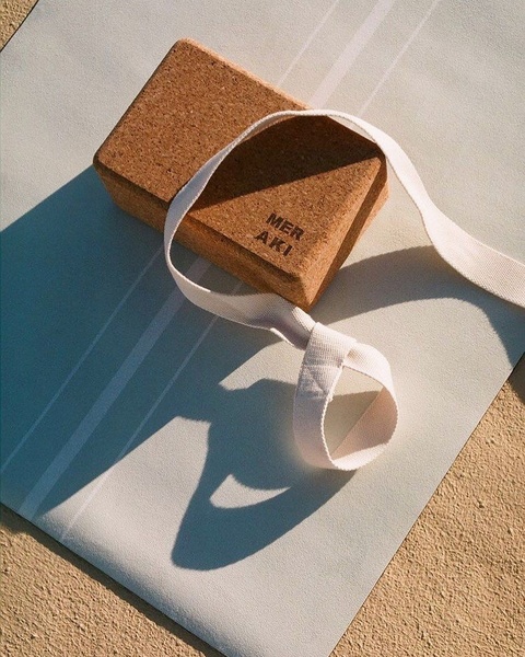 A high-angle, sunlit studio shot of a branded cork yoga block with a white cotton strap resting on a light-colored, striped yoga mat.